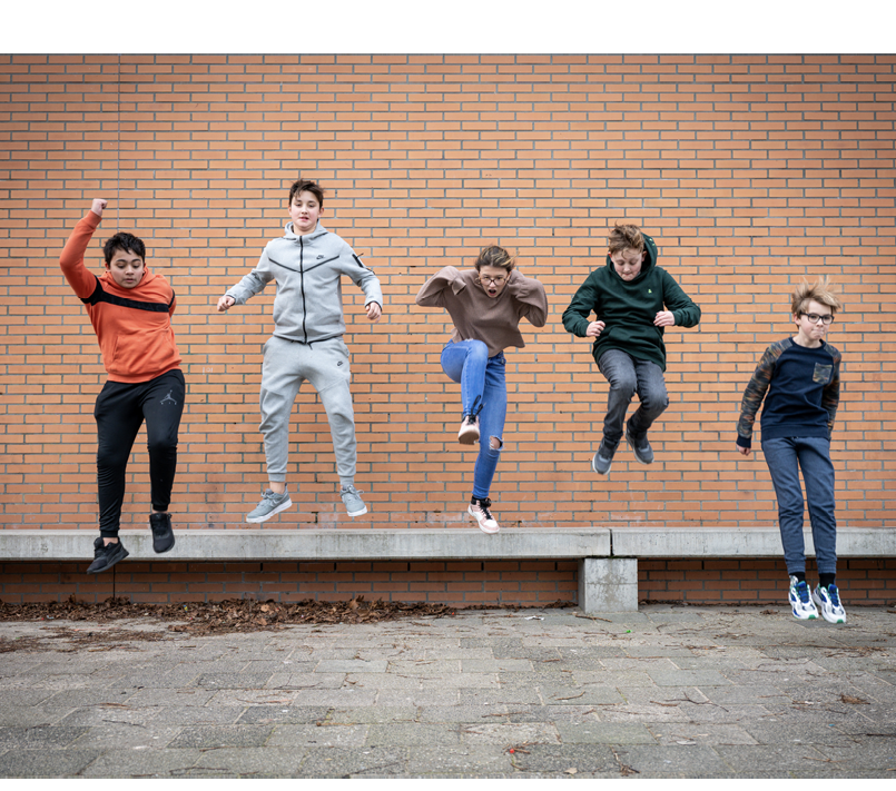 Vijf kinderen springen tegelijk van een betonnen bankje voor een bakstenen muur met verschillende sportieve kleding en energieke houdingen.

Beschrijving gegenereerd door AI